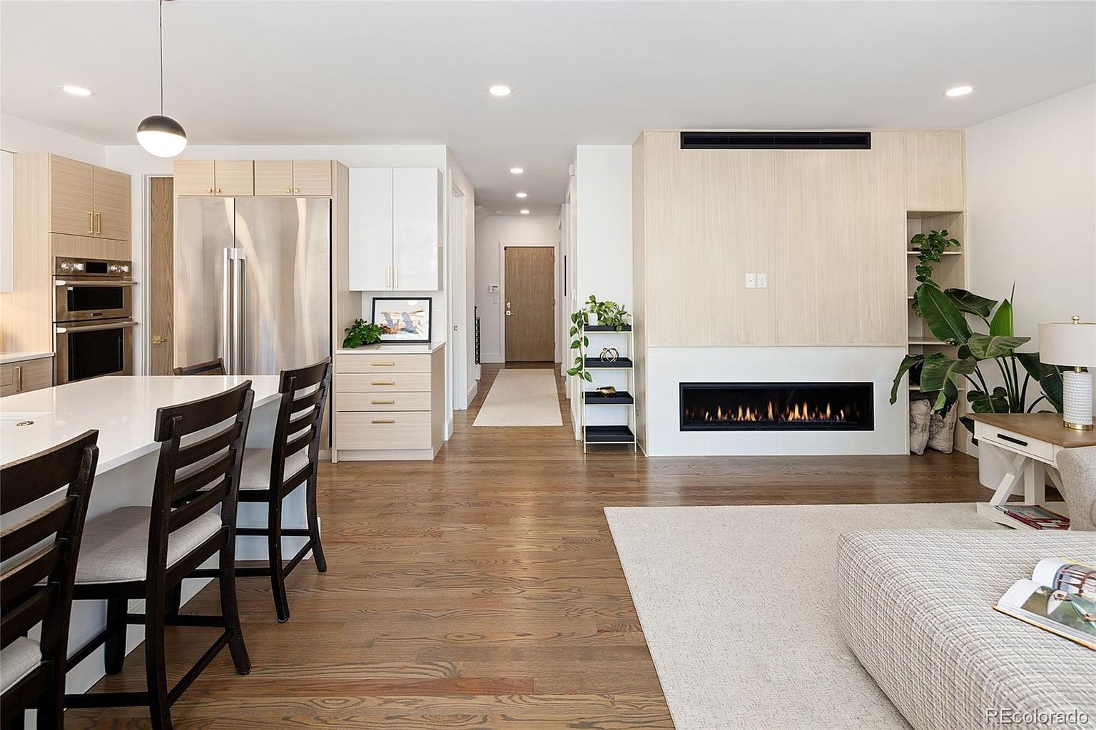 This interior shot showcases a modern living room with a linear fireplace as a focal point, complemented by light wood paneling and built-in shelving. The open-concept design flows into the kitchen area, featuring a large island with bar seating and stainless steel appliances. Hardwood floors and a neutral color palette create a warm and inviting atmosphere.