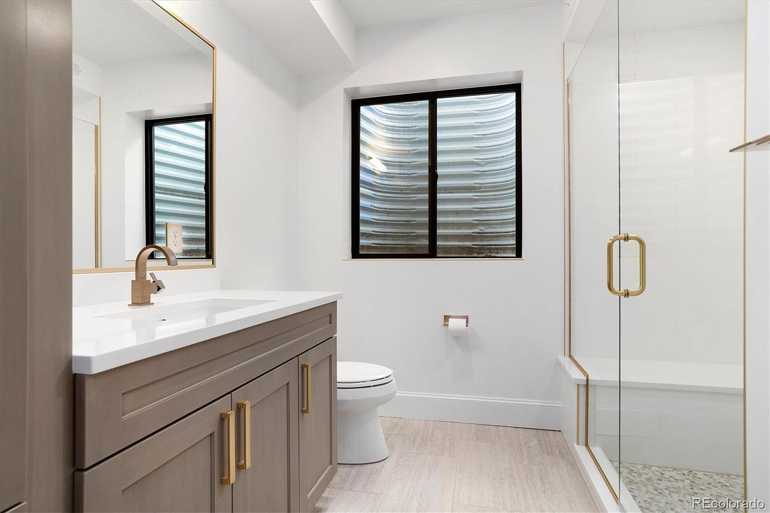 This is a well-lit guest bathroom featuring a modern design. The vanity has a light brown cabinet with gold hardware, a white countertop, and a gold faucet. A black-framed window provides natural light, and a glass-enclosed shower with gold trim adds a touch of luxury.