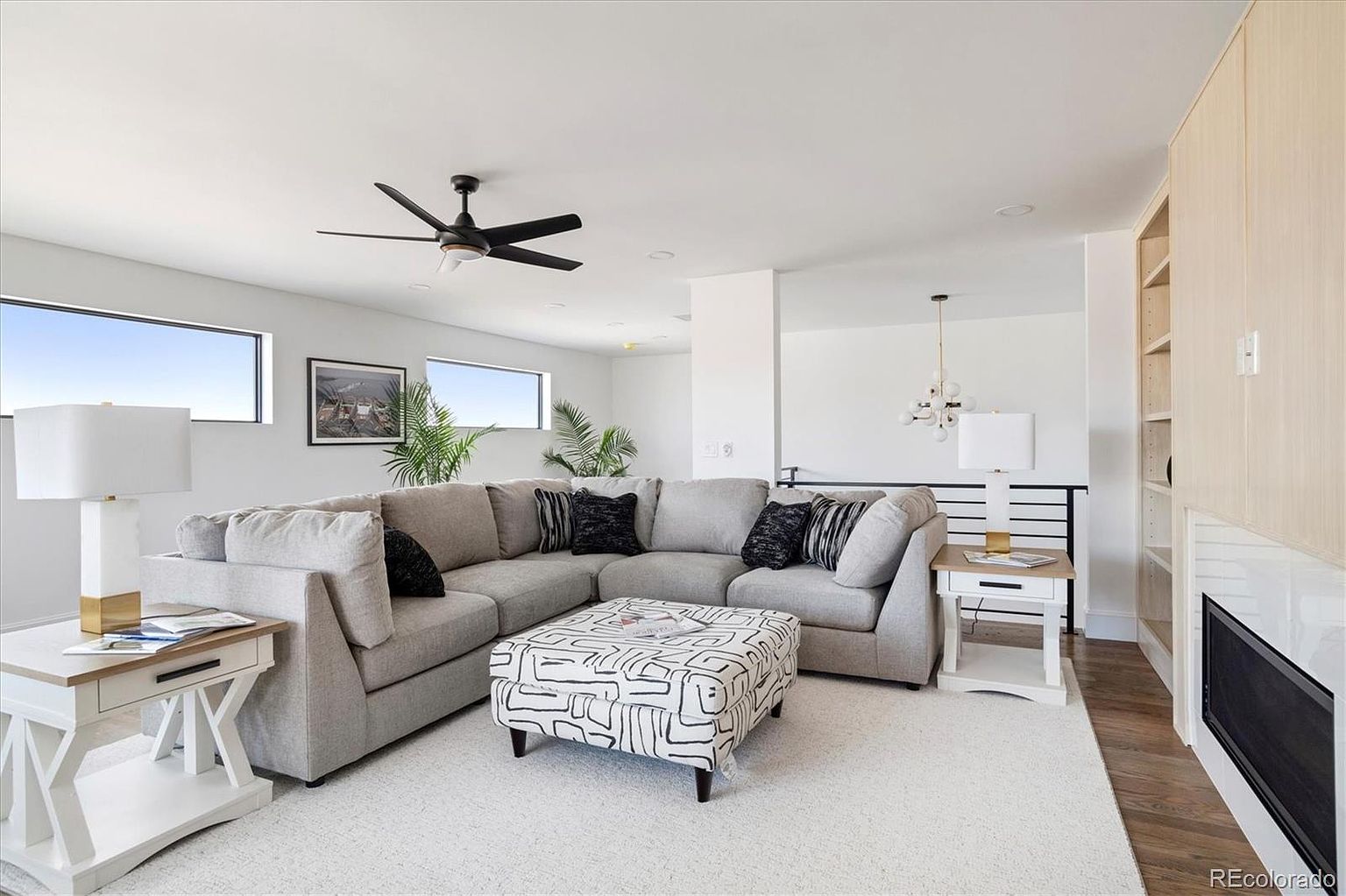 This is a bright and airy living room featuring a large, comfortable sectional sofa with decorative pillows, a patterned ottoman, and white side tables with lamps. The room has a modern aesthetic with clean lines, neutral colors, and natural light streaming in through the windows. A ceiling fan and a fireplace add to the room's appeal.