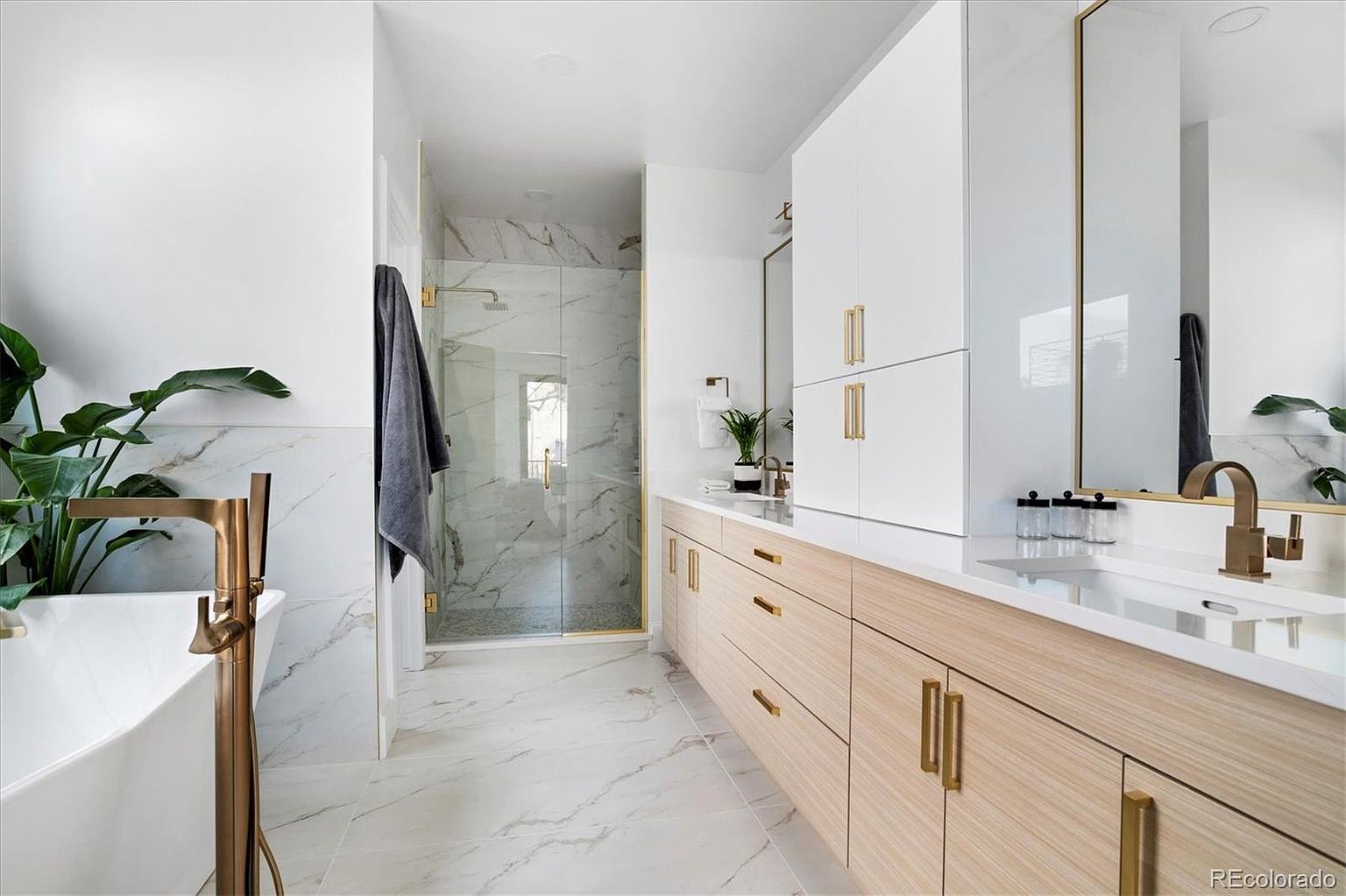 This is a bright and modern primary bathroom featuring marble-look tile flooring and walls, a freestanding tub with a bronze faucet, and a glass-enclosed shower. The vanity has light wood cabinets with bronze hardware, a white countertop, and a matching bronze faucet. White upper cabinets and a large mirror complete the space, creating a luxurious and spa-like atmosphere.