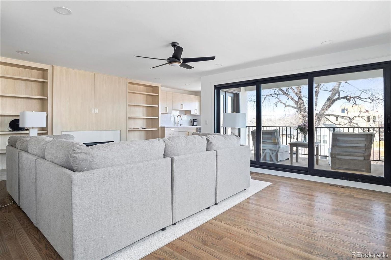 This is a bright and airy living room featuring hardwood floors and a large sectional sofa. Built-in shelving and cabinetry provide ample storage, while a modern ceiling fan adds a touch of style. Sliding glass doors open onto a balcony, offering natural light and outdoor access.