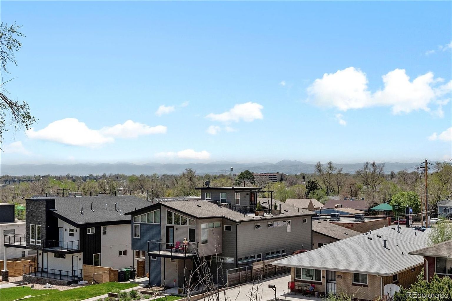 This aerial view showcases a neighborhood with modern homes under a bright, partly cloudy sky. The houses feature contemporary architectural designs, including flat roofs, balconies, and a mix of siding materials. In the background, a mountain range adds depth and scenic appeal, enhancing the property's location.