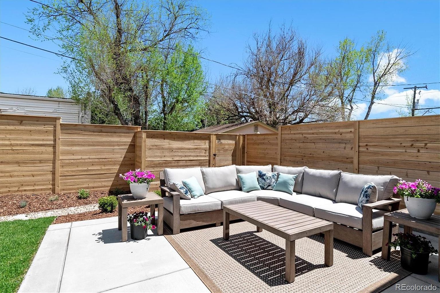 This is a well-staged outdoor patio area, perfect for relaxation and entertaining. The space features a large sectional sofa with neutral cushions and decorative pillows, a wooden coffee table, and side tables adorned with potted flowers. A woven outdoor rug defines the seating area, while a modern wooden fence provides privacy and a clean backdrop.