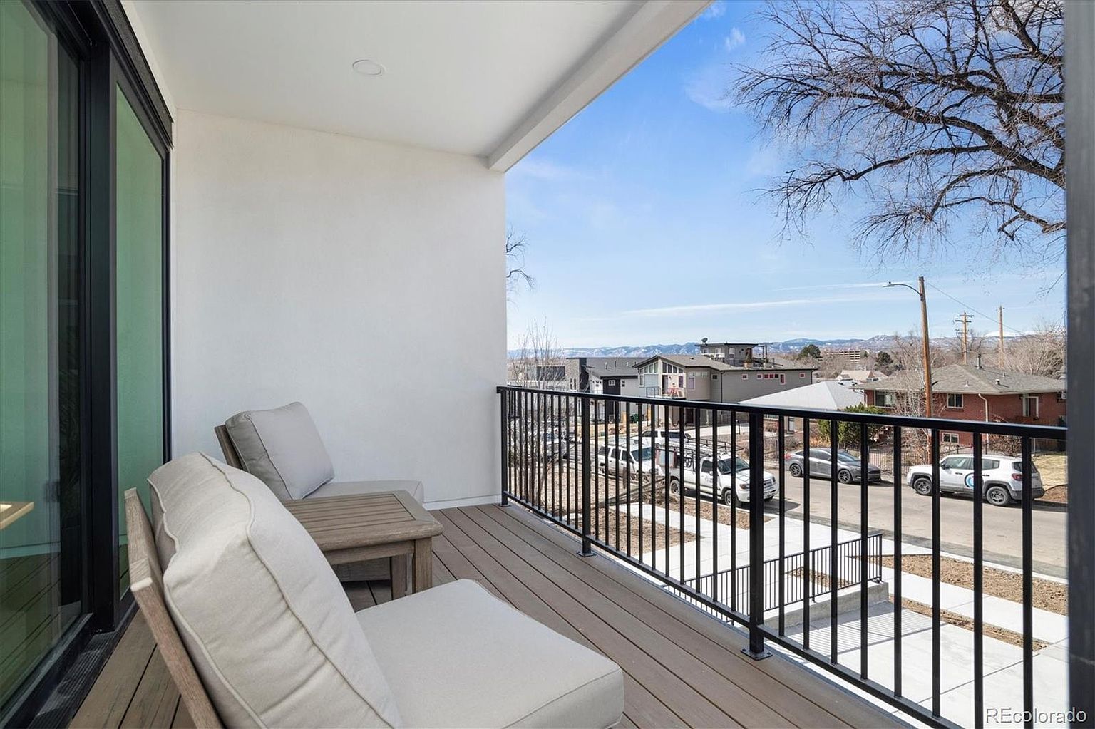 This image showcases a modern balcony with a comfortable seating area. The balcony features a wooden deck, a black metal railing, and a white exterior wall. The seating area includes a cushioned bench and a small side table, creating an inviting outdoor space with a view of the neighborhood and distant mountains.