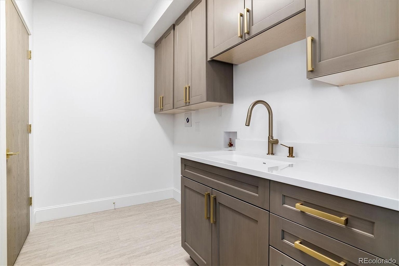 This interior shot showcases a well-organized laundry room featuring stylish gray cabinetry with gold hardware, a white countertop with an integrated sink, and a modern faucet. The room is bright and clean, with light-colored tile flooring and white walls, creating a functional and aesthetically pleasing space.