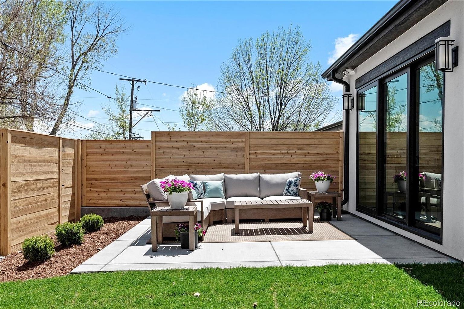 This image showcases a beautifully designed outdoor patio area. The space features a modern wooden sectional sofa with neutral cushions and decorative pillows, complemented by matching side tables and a coffee table. A wooden fence provides privacy, while a well-maintained lawn and landscaping add to the inviting atmosphere, making it an ideal space for relaxation and entertaining.
