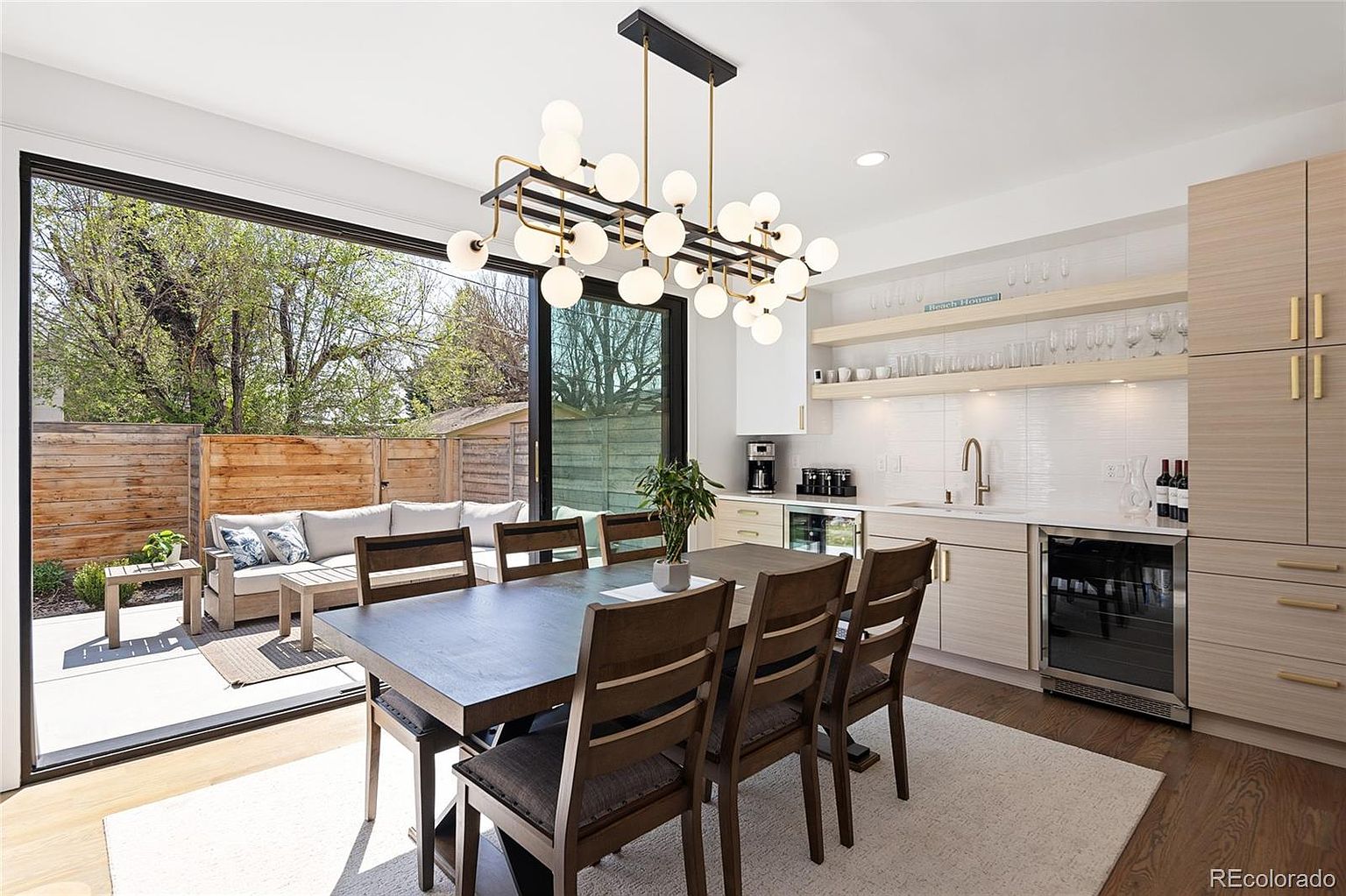 This is an interior shot of a dining room with a modern aesthetic. The room features a large dining table with six chairs, a contemporary chandelier, and a bar area with wine storage. Sliding glass doors open to an outdoor patio area, creating a seamless indoor-outdoor living space.