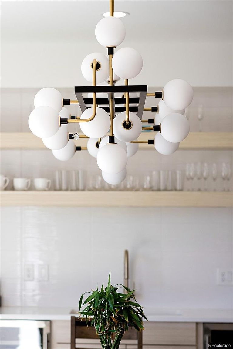 This image showcases a modern kitchen interior, focusing on a stylish chandelier with spherical white shades and a black and gold frame. The background features light wood shelves displaying glassware and dishware, complemented by white tiled walls and countertops. A green plant adds a touch of nature to the scene, creating a bright and inviting atmosphere.