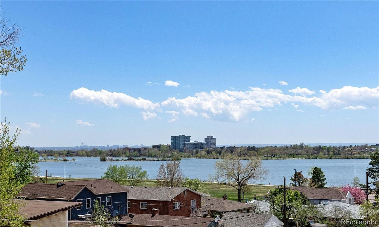 This aerial view showcases a residential area with houses overlooking a serene lake. The cityscape is visible in the distance, framed by a clear blue sky with scattered clouds. The scene provides a sense of tranquility and highlights the desirable location near the water and city amenities.