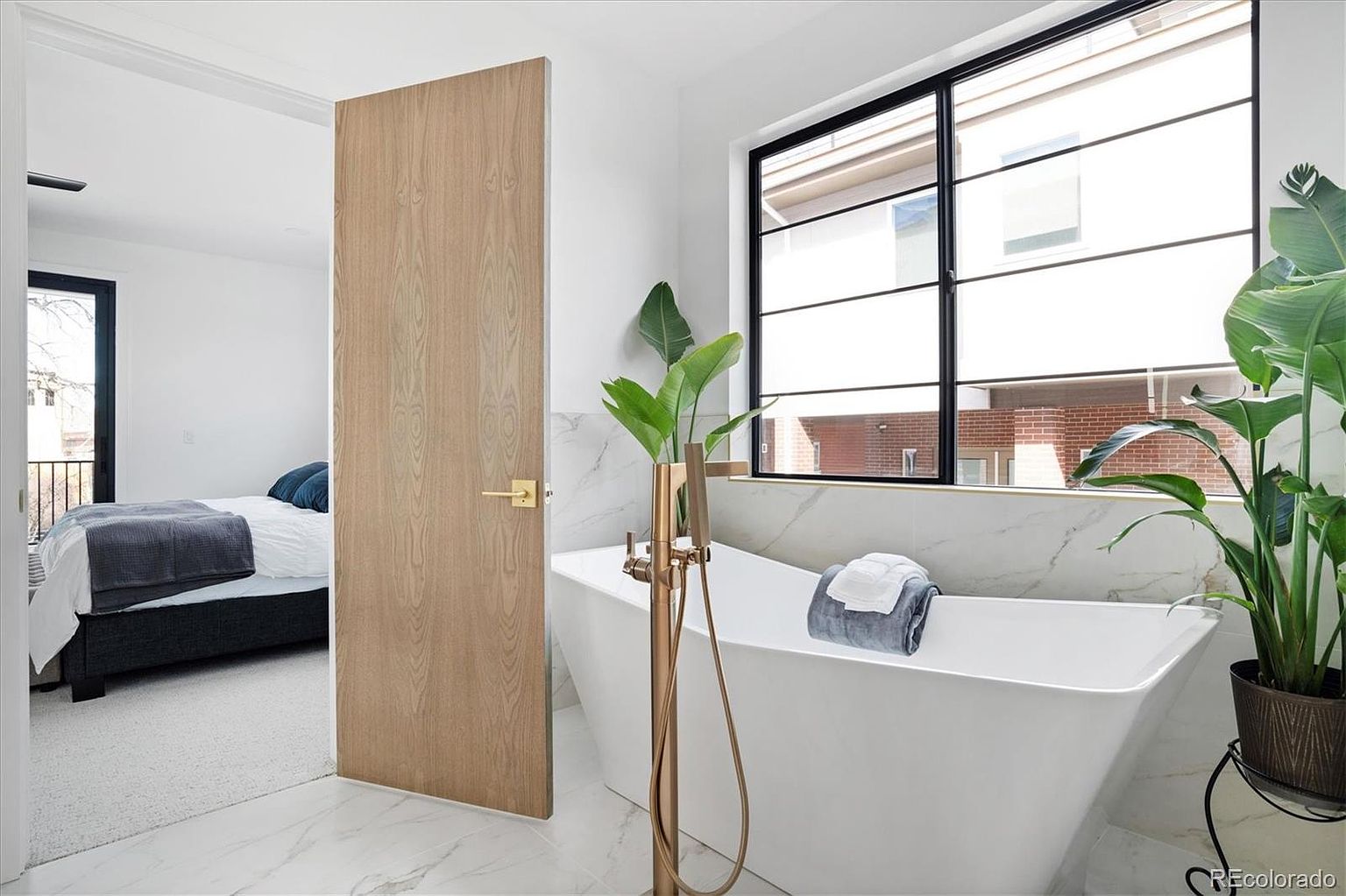 This is a bright and modern primary bathroom featuring a freestanding white bathtub with a gold faucet and a large window providing natural light. The walls and floor are covered in marble, and a potted plant adds a touch of nature to the space. A bedroom is visible through an open doorway, suggesting an en-suite layout.