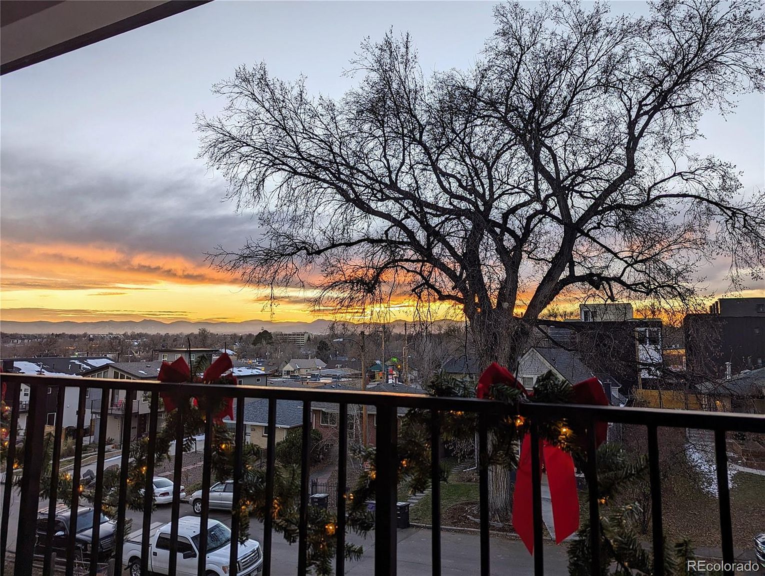 This image showcases a balcony view at sunset, framed by a black metal railing adorned with festive wreaths and red bows. The cityscape stretches into the distance, with mountains silhouetted against the vibrant orange and yellow sky. A large, bare tree dominates the foreground, adding a touch of natural beauty to the urban scene.