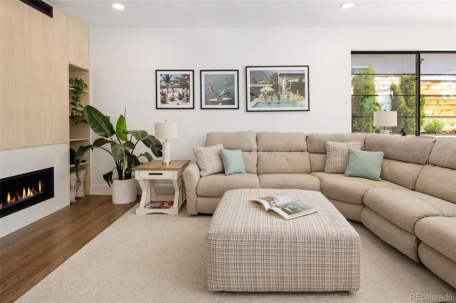 This is an interior shot of a living room featuring a large, beige sectional sofa with throw pillows, a patterned ottoman, and three framed art pieces hanging above the sofa. A modern fireplace is built into a light wood-paneled wall, and a large potted plant sits next to a side table with a lamp. The room is well-lit and appears comfortable and inviting.