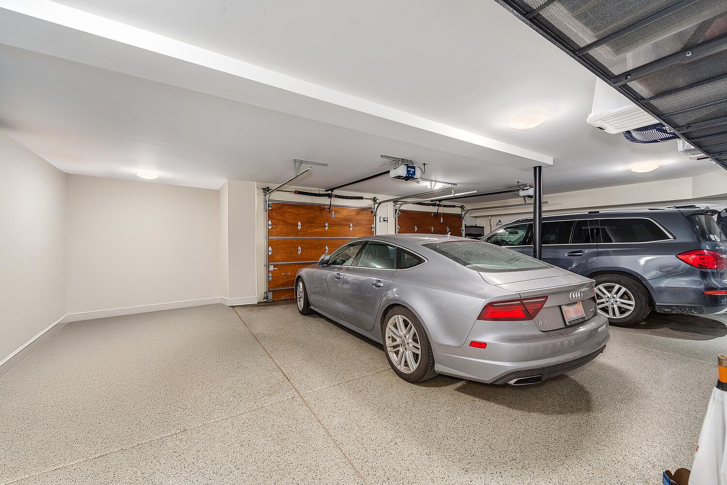 The image showcases a well-organized garage featuring two cars, a silver sedan and a dark SUV. The garage has two wooden doors with automatic openers, a light-colored epoxy floor, and white walls and ceiling. Overhead storage racks are visible, adding to the functionality of the space.