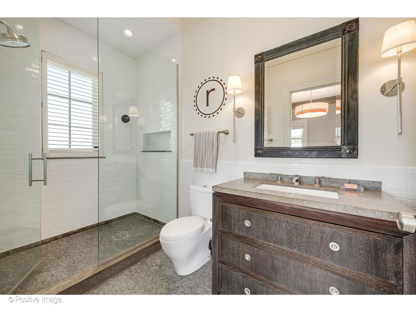 This is a well-lit primary bathroom featuring a glass-enclosed shower with gray tiled floors and white subway tile walls. The vanity has a dark wood finish with a light-colored countertop, complemented by a framed mirror and sconce lighting. A toilet is positioned next to the shower, and the overall design is clean and modern.