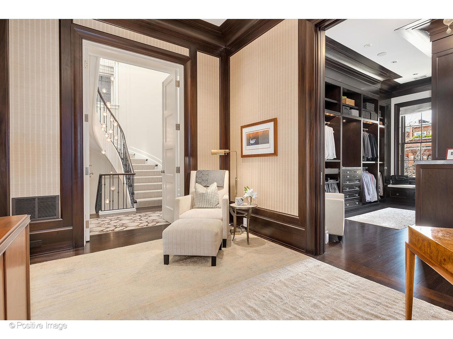 This is a view of a primary bedroom that leads into a walk-in closet and a staircase. The bedroom features dark wood trim, a neutral color palette, and a comfortable armchair with an ottoman. The walk-in closet is well-organized with custom shelving and drawers, while the staircase has a wrought iron railing and leads to another level.