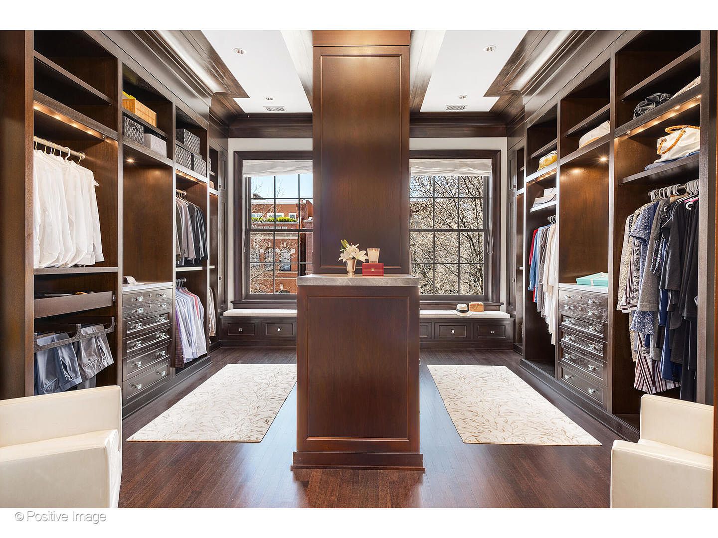 This is a luxurious walk-in closet featuring dark wood cabinetry, ample shelving, and hanging space. A central island with a marble countertop provides additional storage and display space. Natural light streams in through two windows, enhancing the elegant and organized feel of the closet.
