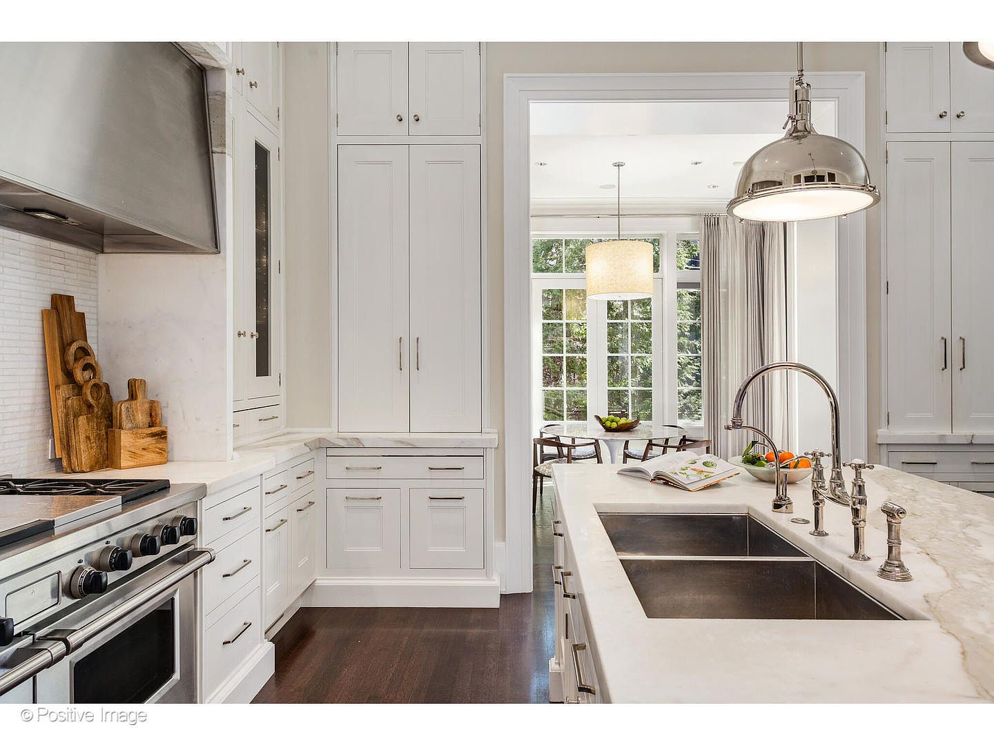 This is a bright and elegant kitchen featuring white cabinetry, marble countertops, and stainless steel appliances. A large island with a sink and faucet is prominently displayed, with a view into an adjacent dining area through a wide doorway. The dark hardwood floors provide a nice contrast to the light color palette, creating a sophisticated and inviting space.