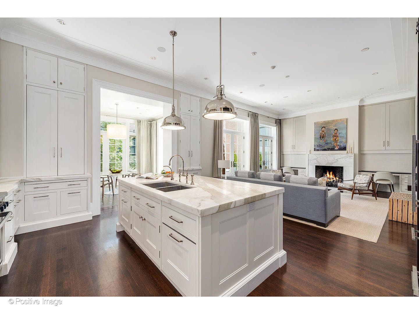This is a bright and open kitchen and living space featuring a large kitchen island with a marble countertop and stainless steel sink. The kitchen has white cabinetry and dark hardwood floors, seamlessly flowing into a living area with a fireplace and comfortable seating. The perspective is from a wide angle, showcasing the spaciousness and connectivity of the rooms.