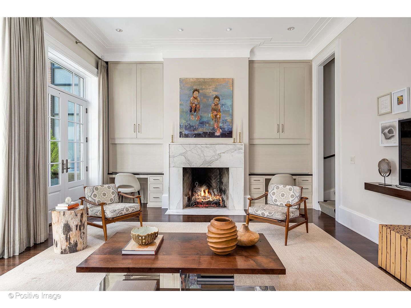 This is an interior shot of a living room featuring a fireplace with a marble surround and a painting above it. Two armchairs face the fireplace, flanking a dark wood coffee table with decorative vases. The room has built-in cabinets, a large window with curtains, and a neutral-toned rug, creating a cozy and elegant atmosphere.