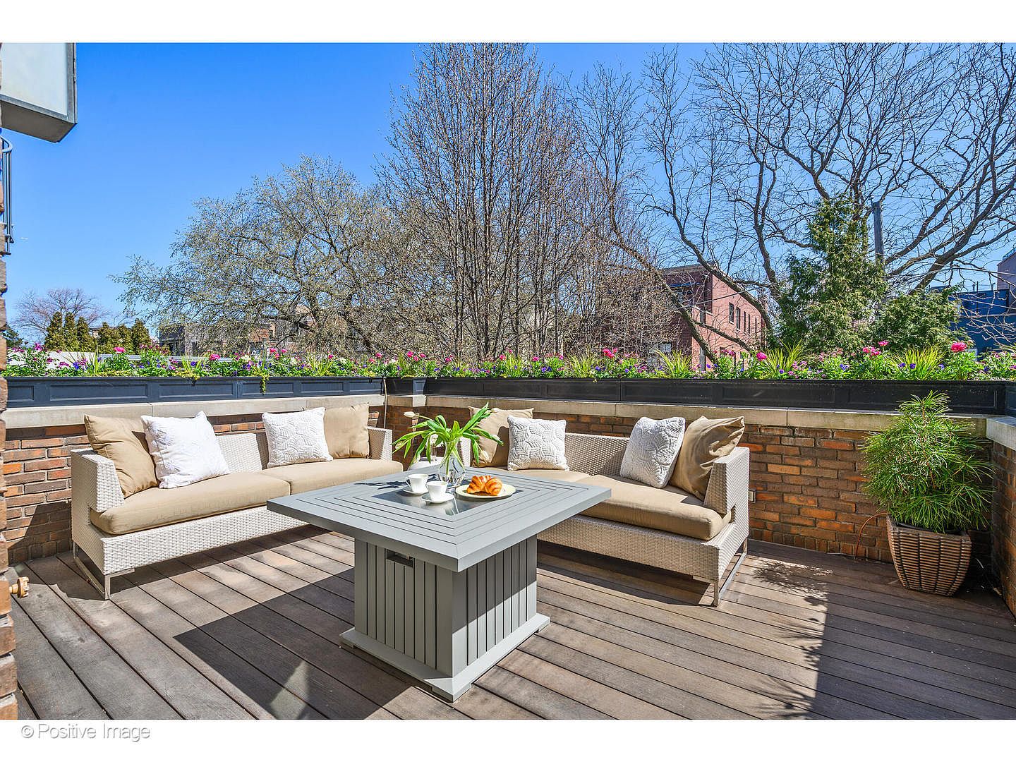 This image showcases a beautifully arranged outdoor patio or balcony area. The space features comfortable wicker seating with neutral-toned cushions and decorative pillows, centered around a modern gray coffee table set with breakfast items. The wooden deck flooring and brick railing with flower boxes create a cozy and inviting atmosphere, perfect for relaxation or entertaining.