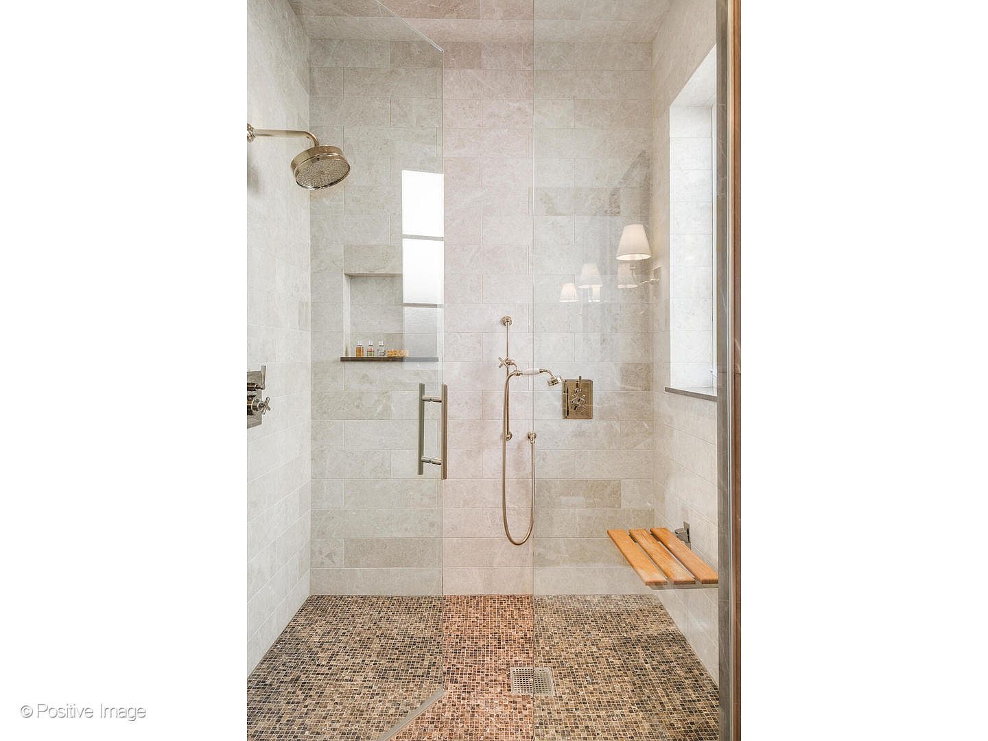 This is a well-lit primary bathroom showcasing a modern shower. The shower features neutral-toned tiled walls, a mosaic tile floor, and a glass enclosure. A built-in niche provides storage, and a wooden bench offers seating, creating a spa-like atmosphere.