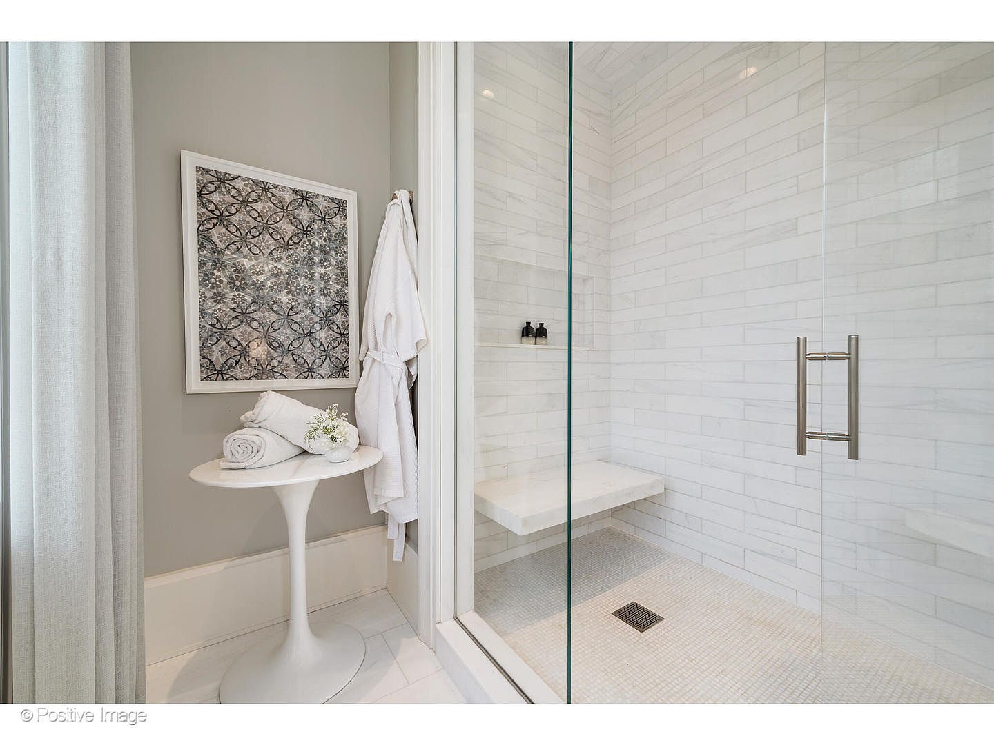 This is a bright and modern primary bathroom featuring a glass-enclosed shower with a built-in bench and a sleek, minimalist design. The shower walls are tiled with horizontal white tiles, and the floor is covered in a mosaic pattern. A white tulip table with rolled towels and a framed artwork adds a touch of elegance to the space.