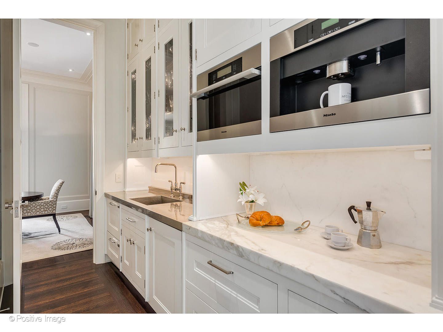 This image showcases a well-appointed kitchen area with white cabinetry, stainless steel appliances, and marble countertops. A built-in coffee machine and microwave are featured above the counter, which displays croissants, cups, and a moka pot. The space is illuminated by under-cabinet lighting, creating a warm and inviting atmosphere.