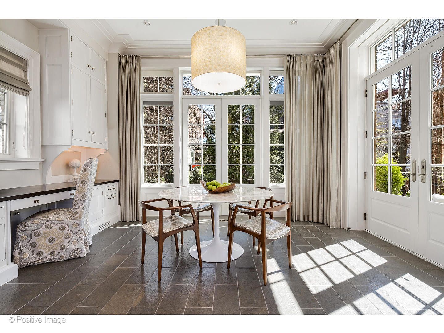 This is an interior shot of a sunlit dining room. The room features a round marble-top table with four wooden chairs, a large drum pendant light, and a wall of windows with neutral-toned curtains. The floor is tiled with large gray stones, and there is a built-in desk area with white cabinets on the left side of the image.