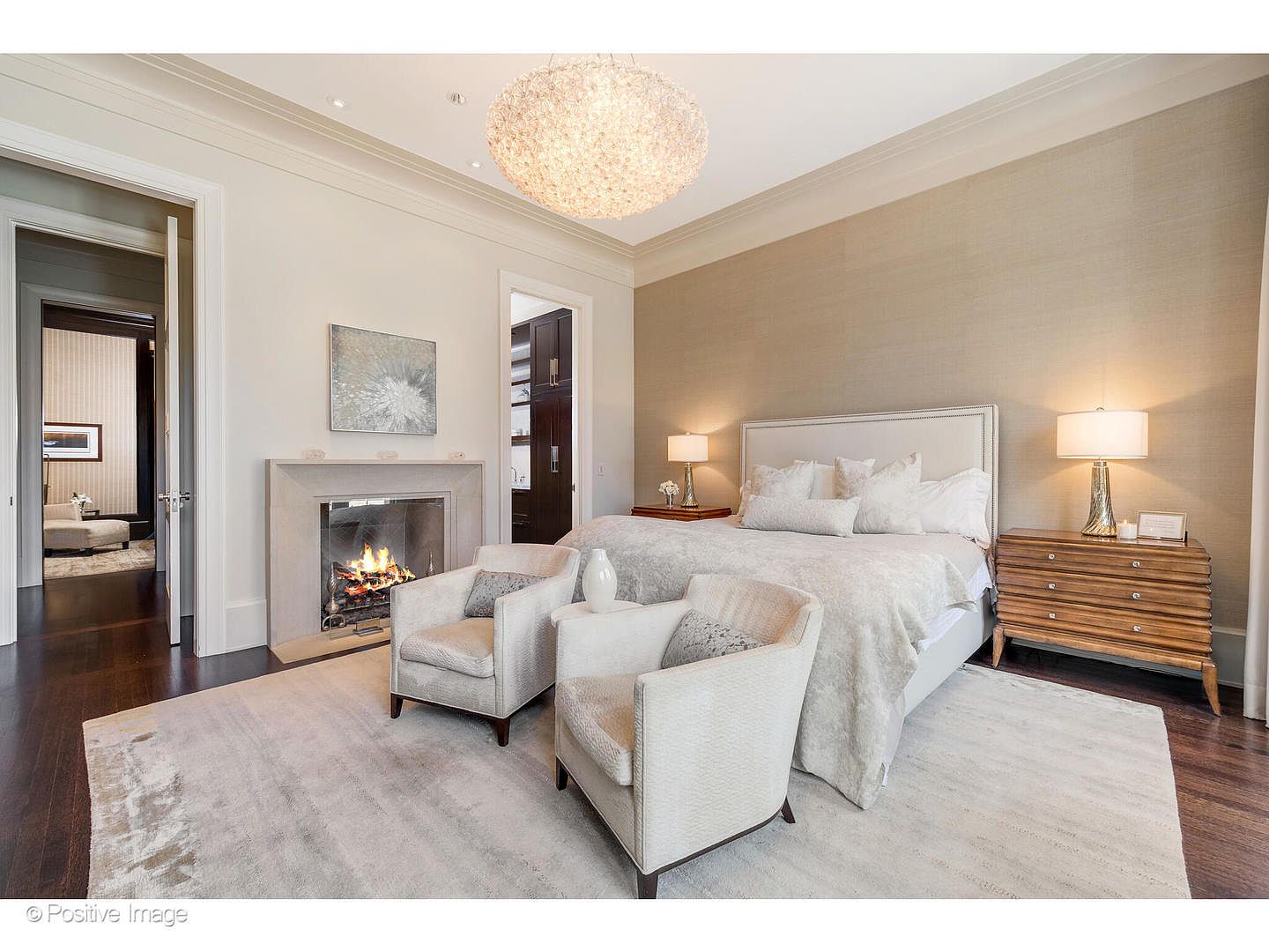 This is a primary bedroom featuring a neutral color palette with a large bed, two armchairs, and a fireplace. The room is well-lit with a decorative chandelier and bedside lamps, creating a warm and inviting atmosphere. The hardwood floors and area rug add to the luxurious feel of the space.
