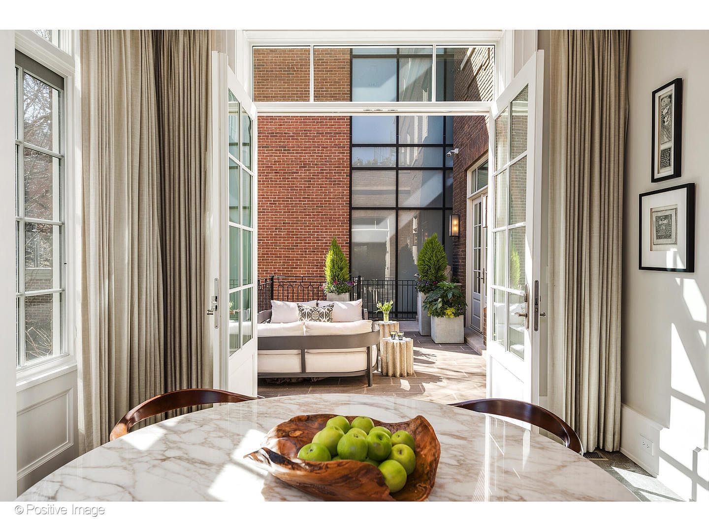 This image showcases a dining room that seamlessly transitions to an outdoor patio. The dining area features a round marble table with a wooden bowl of green apples as a centerpiece. Through the open French doors, a cozy outdoor seating area with a sofa and potted plants is visible, creating an inviting indoor-outdoor living space.
