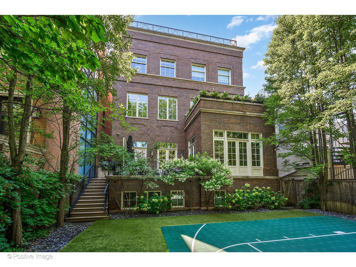 This is a rear view of a multi-story brick building, likely a residential property, showcasing a well-maintained backyard. The yard features a combination of green lawn, a basketball court, and lush landscaping with trees and flowering bushes. A staircase leads up to a higher level of the building, suggesting outdoor living spaces or balconies.