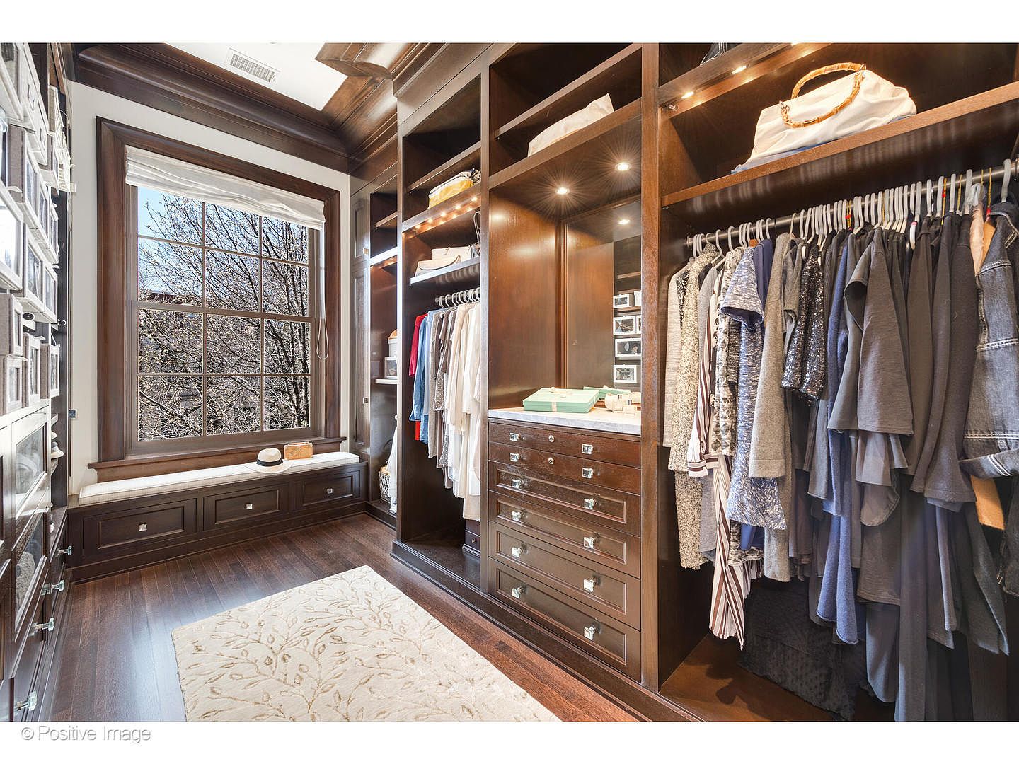 This is a well-organized walk-in closet featuring custom dark wood cabinetry and shelving. The closet includes hanging rods filled with clothing, drawers with decorative hardware, and a window seat with natural light. A patterned area rug adds a touch of elegance to the hardwood flooring.