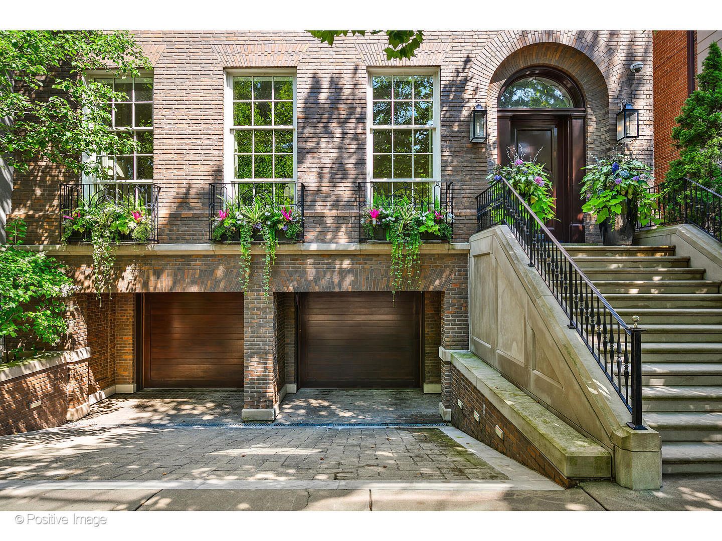 This is a front exterior view of a brick townhouse featuring two garage doors, a set of stairs leading to the main entrance, and multiple windows with flower boxes. The architecture is traditional with an arched doorway and wrought iron railings, creating a sophisticated and inviting curb appeal. The landscaping is well-maintained, adding to the overall charm of the property.