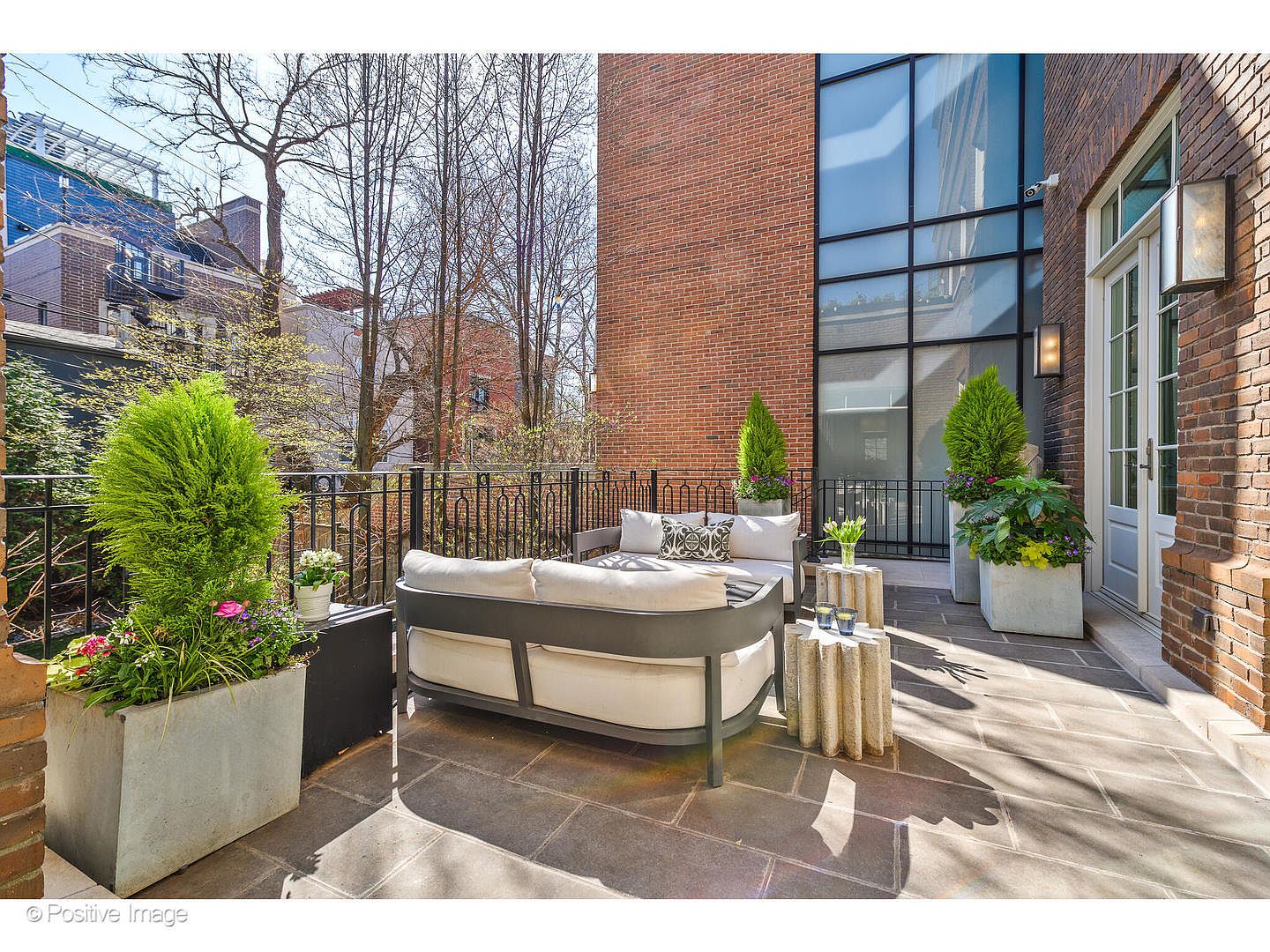 This image showcases a stylish outdoor patio area, featuring a modern sofa with light cushions, complemented by decorative pillows and small side tables. The patio is adorned with potted plants and trees, adding a touch of greenery and elegance. The space is paved with stone tiles and is enclosed by a black metal railing, offering a private and sophisticated outdoor living area.
