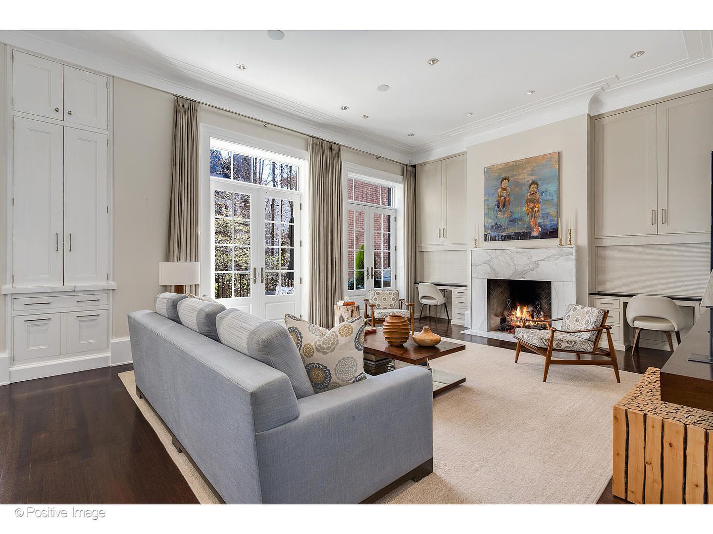 This is a well-lit living room featuring a light gray sofa, a fireplace with a marble surround, and built-in cabinetry. The room has a neutral color palette with dark hardwood floors and a large area rug. French doors lead to the outside, and artwork hangs above the fireplace, creating a sophisticated and inviting atmosphere.