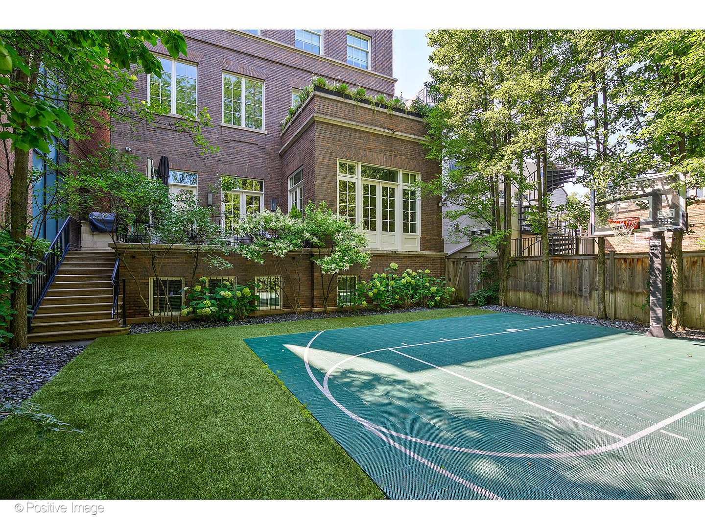 This exterior shot showcases a well-maintained backyard featuring a basketball court and lush green lawn. The brick building provides a sophisticated backdrop, complemented by mature trees and manicured landscaping. A staircase leads to an upper level, adding architectural interest and suggesting outdoor living spaces.