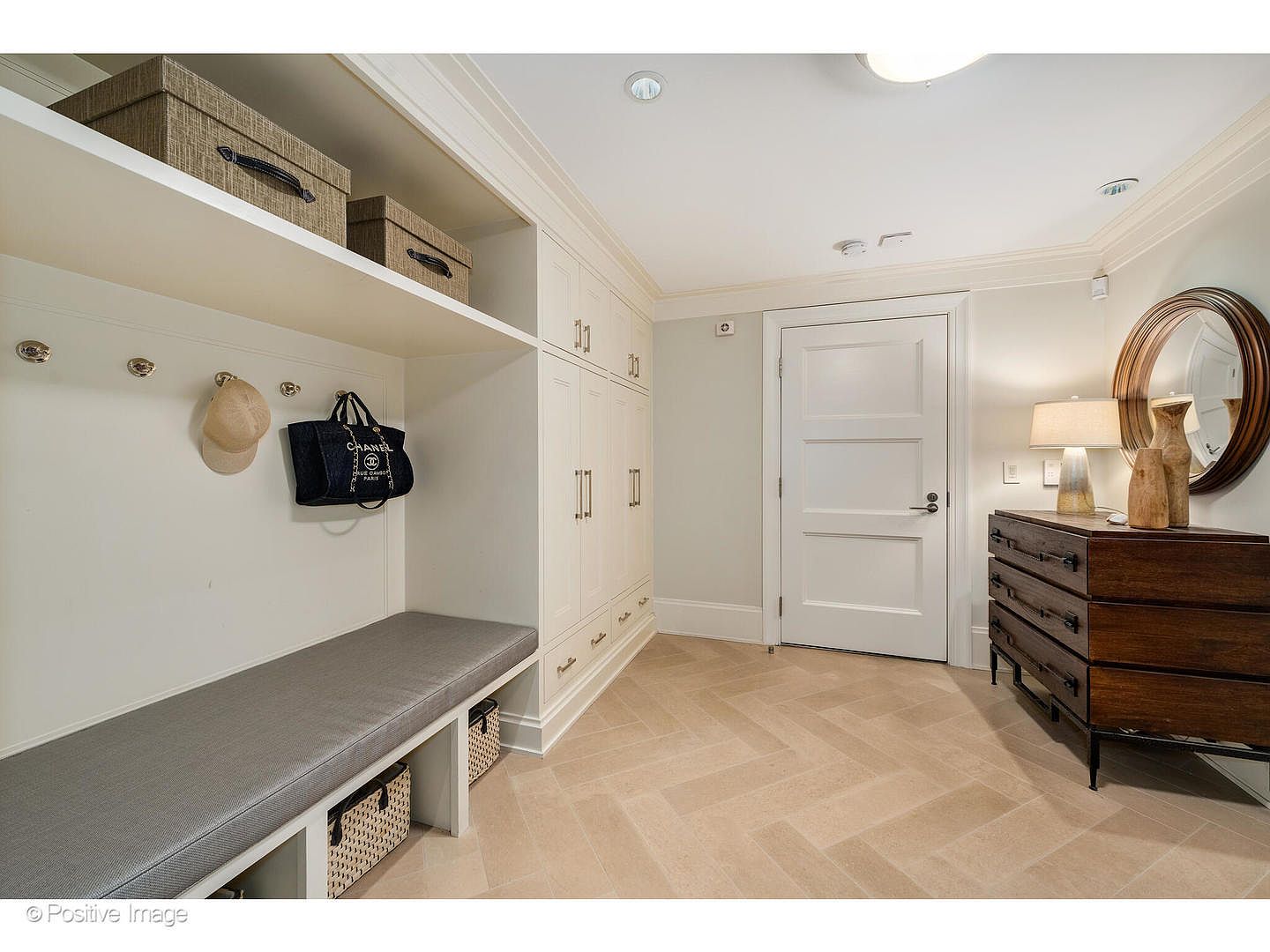 This interior shot showcases a well-organized mudroom or hallway area. It features built-in storage with a bench seat, overhead shelving, and enclosed cabinets, all in a neutral color palette. A dark wood dresser with a round mirror above it adds a touch of elegance, while the herringbone-patterned tile flooring contributes to the room's sophisticated design.