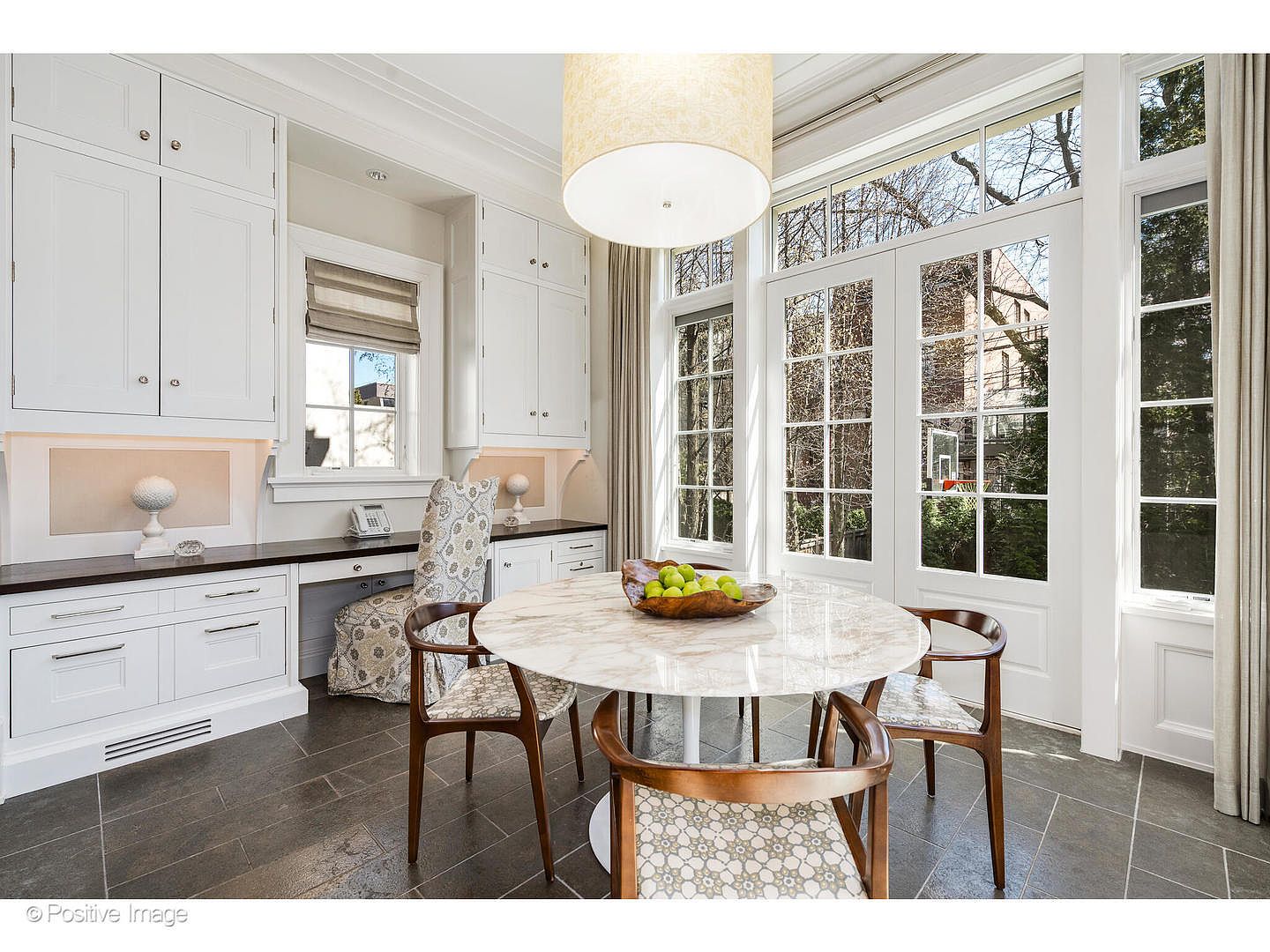 This is an interior shot of a bright and airy dining room. The room features a round marble table surrounded by four chairs with patterned upholstery and wooden frames. Large windows with white frames and a glass door provide ample natural light, complemented by a modern drum pendant light fixture. Built-in white cabinets and a desk area add functionality to the space, while the dark gray tile flooring provides a sophisticated contrast.