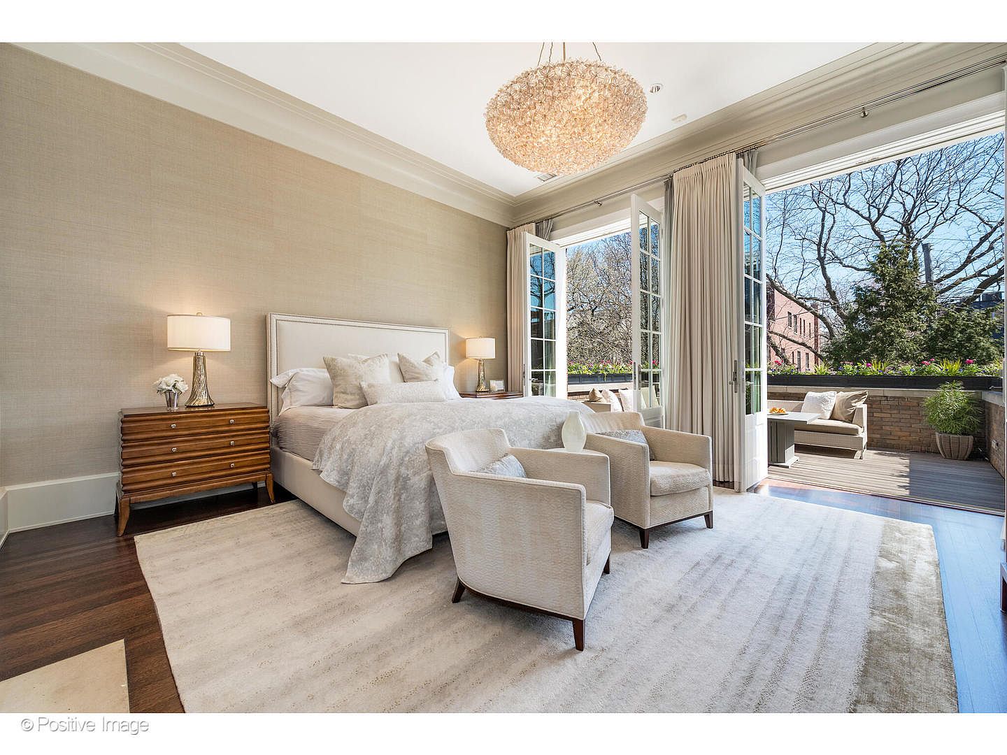 This is a luxurious primary bedroom featuring a king-size bed with white linens and decorative pillows. The room is well-lit with a statement chandelier and natural light streaming in from the open French doors leading to a balcony. Two armchairs sit on a large area rug, creating a cozy seating area within the bedroom.