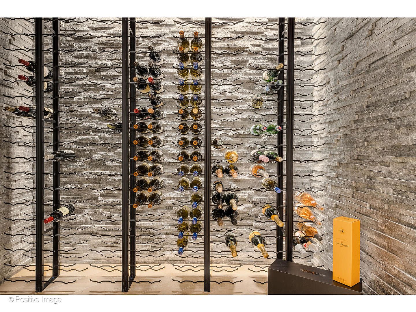 This image showcases a sophisticated wine cellar with a modern design. The wine bottles are neatly arranged in a black metal racking system against a textured stone wall, creating an elegant display. A Veuve Clicquot champagne box adds a touch of luxury, suggesting a high-end and well-curated collection.