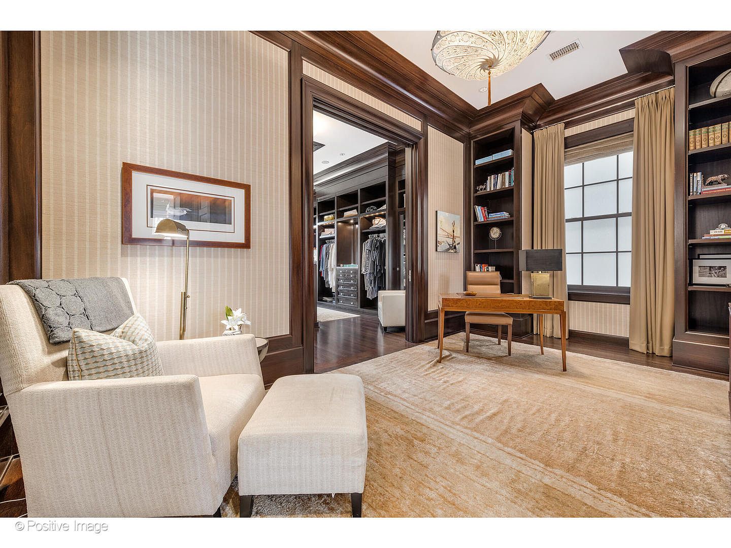 This is a well-appointed home office featuring dark wood trim, built-in bookshelves, and a comfortable seating area. A wooden desk sits in front of a window with natural light, and a doorway leads to a walk-in closet. The room exudes a sense of sophistication and functionality, perfect for a professional working from home.