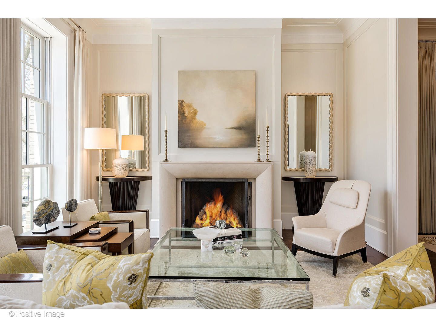 This is an elegant living room featuring a fireplace as the focal point, flanked by matching mirrors and console tables. The room is decorated in neutral tones with pops of yellow in the throw pillows, creating a warm and inviting atmosphere. A glass coffee table sits in the center, and the overall style is sophisticated and luxurious.