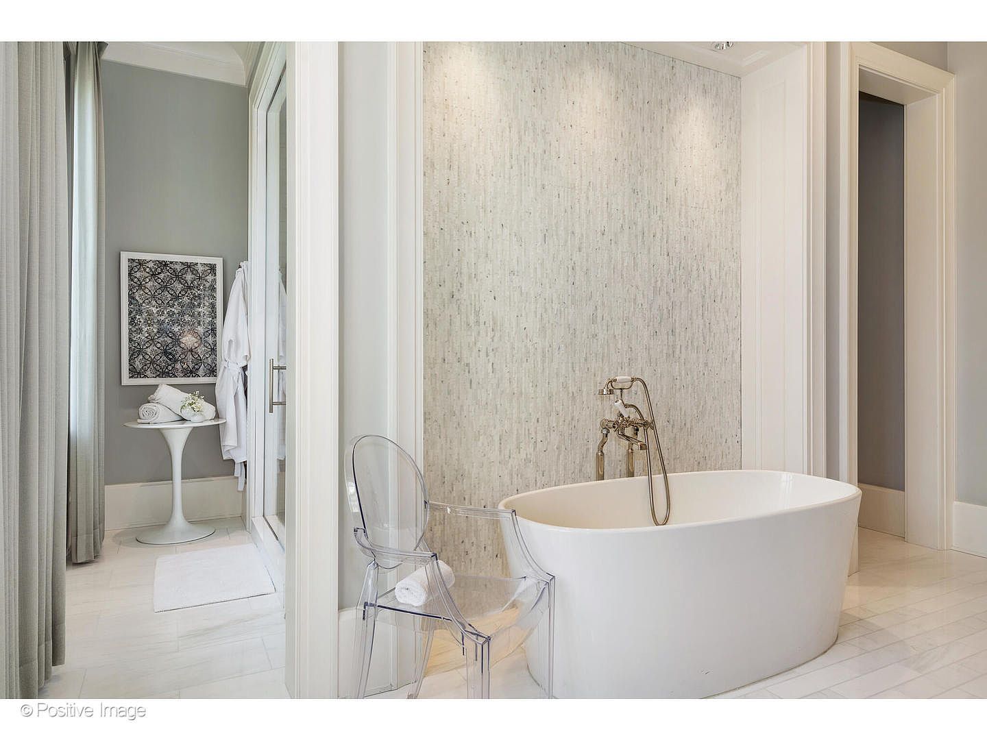 This is a bright and elegant primary bathroom featuring a freestanding white bathtub with a vintage-style faucet. The walls are adorned with textured, light-colored tiles, and a clear acrylic chair sits nearby. The space is well-lit and appears luxurious, with a glimpse into an adjacent area with a vanity and artwork.