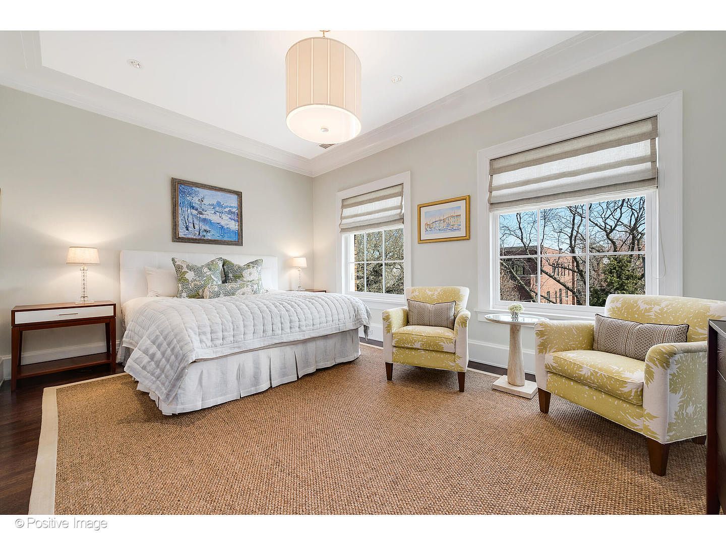 This is a well-lit primary bedroom featuring a large bed with a white quilted comforter and decorative pillows. Two yellow armchairs flank a small side table near the windows, which are adorned with neutral-toned blinds. The room is carpeted with a textured rug, and artwork hangs above the bed and near the windows, creating a serene and inviting atmosphere.
