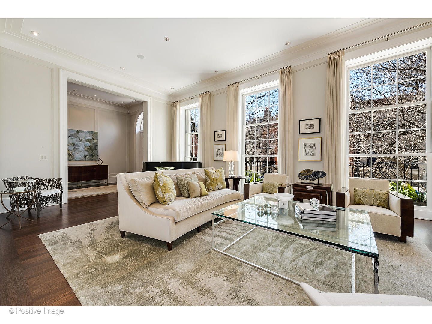 This is a bright and elegant living room featuring large windows that provide ample natural light. The room is furnished with a comfortable sofa, armchairs, and a glass coffee table, all set on a neutral-toned rug. The space is decorated with artwork and accessories, creating a sophisticated and inviting atmosphere.
