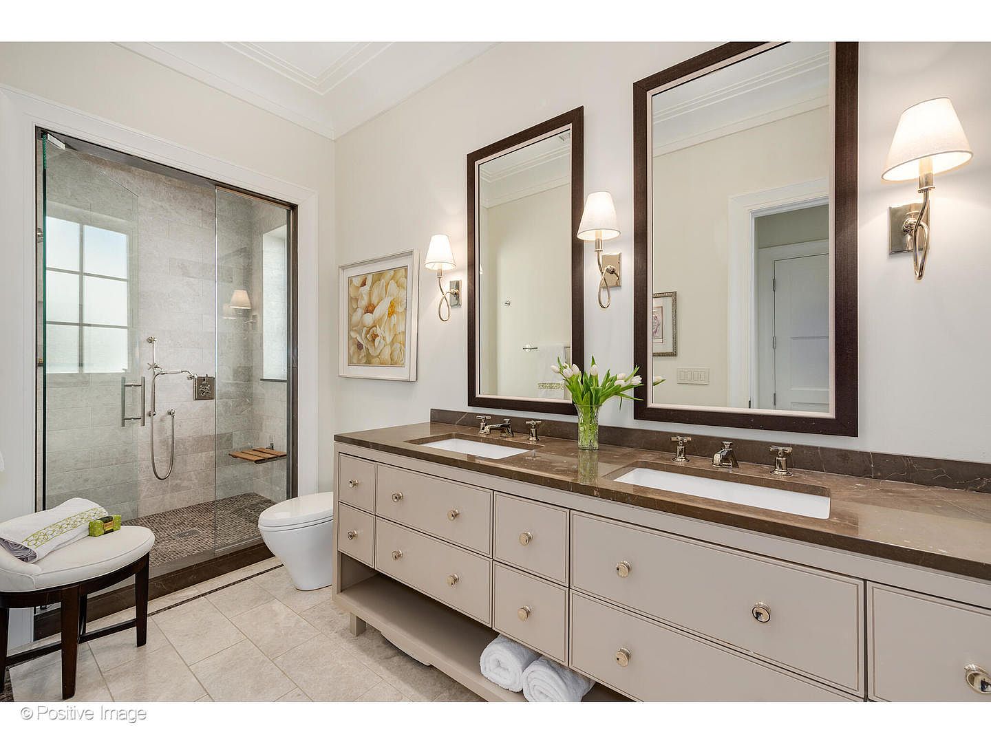 This is a well-lit primary bathroom featuring a double vanity with a brown countertop and beige cabinetry. Two framed mirrors hang above the sinks, each flanked by sconce lighting. A glass-enclosed shower is visible to the left, and a toilet and stool are also present, creating a luxurious and functional space.
