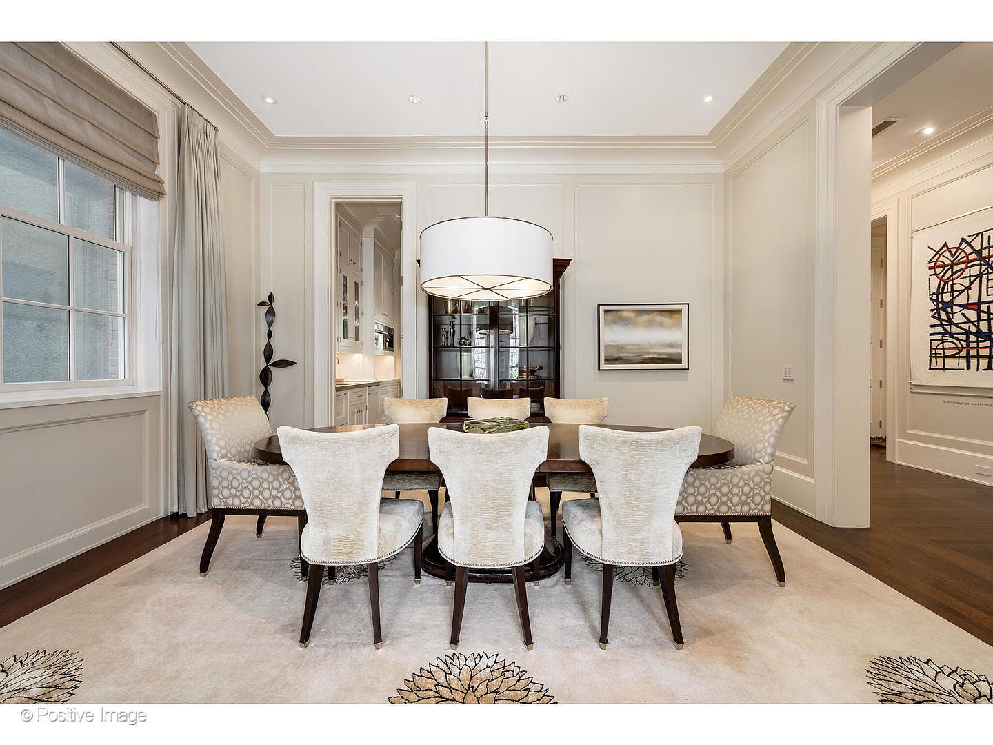 This is an interior shot of a dining room featuring an oval wooden table surrounded by six upholstered chairs. A large, round pendant light hangs above the table, illuminating the space. The room is decorated in neutral tones with elegant molding and trim, and a patterned rug covers the hardwood floor, adding a touch of sophistication.