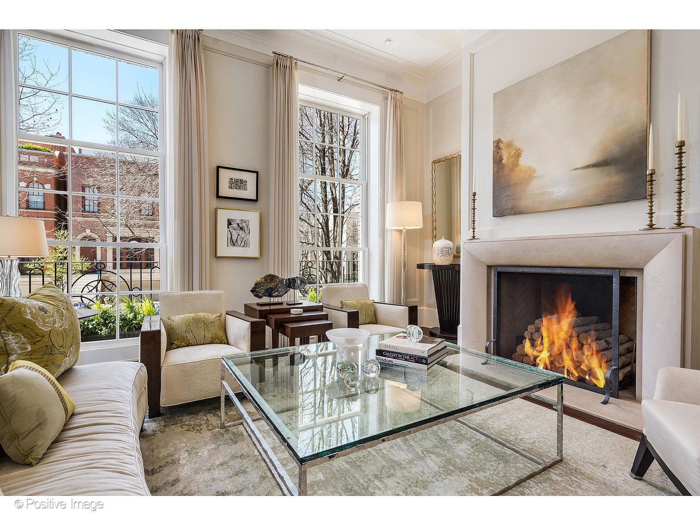 This is an interior shot of a luxurious living room featuring a fireplace with a roaring fire, a glass coffee table, and comfortable seating. Large windows provide ample natural light, and the neutral color palette creates a warm and inviting atmosphere. The room is elegantly decorated with artwork and decorative objects, suggesting a sophisticated and comfortable living space.