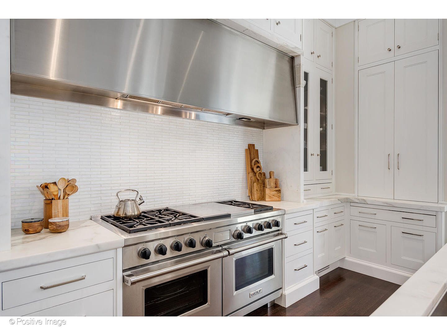 This is a well-lit kitchen featuring white cabinetry, marble countertops, and a stainless steel Wolf range with a large vent hood. The backsplash is a white subway tile, and wooden utensils add a touch of warmth. The dark hardwood floors provide a nice contrast to the light cabinetry, creating a clean and elegant look.