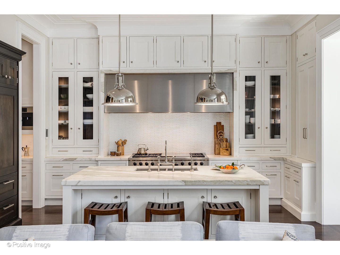This is a bright and spacious kitchen featuring white cabinetry, a large marble island with seating, and stainless steel appliances. The kitchen is well-lit with pendant lighting and natural light, creating a clean and inviting atmosphere. The perspective is from the living room, looking into the kitchen.